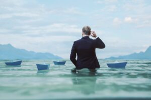 Businessman standing in water scratching head surrounded by paper boats, insolvency concept