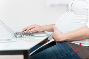 Pregnant woman holding tummy sitting at laptop, maternity leave concept
