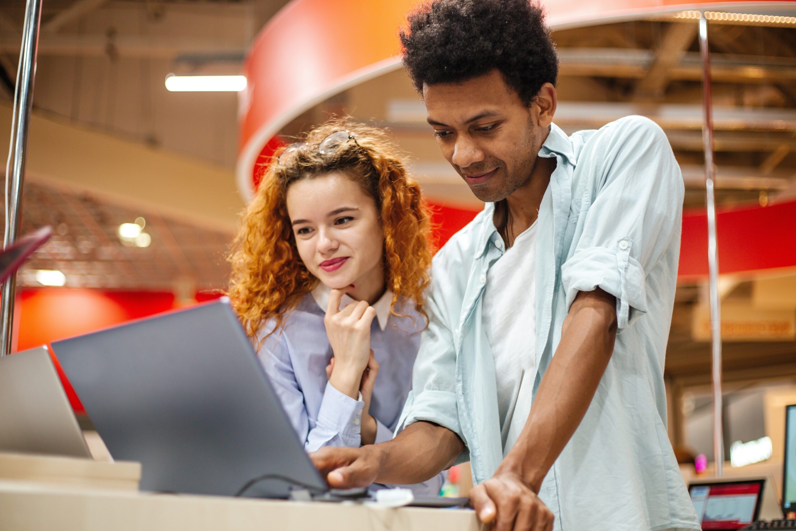 Red-haired standing beside dark-skinned man peering at laptops, buying-in tech concept