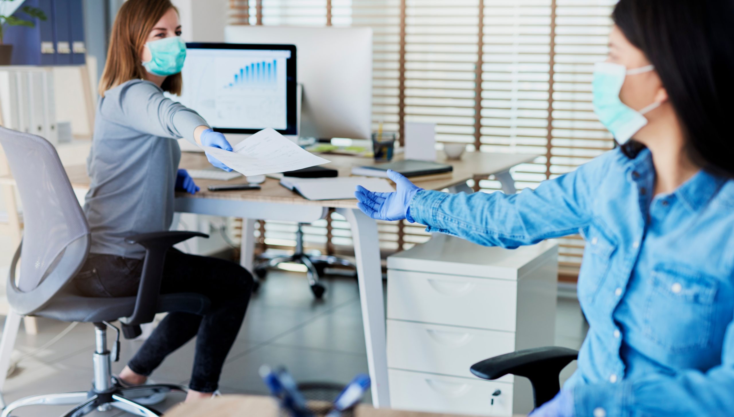Two women handing across a piece of paper wearing facemasks, return to work concept