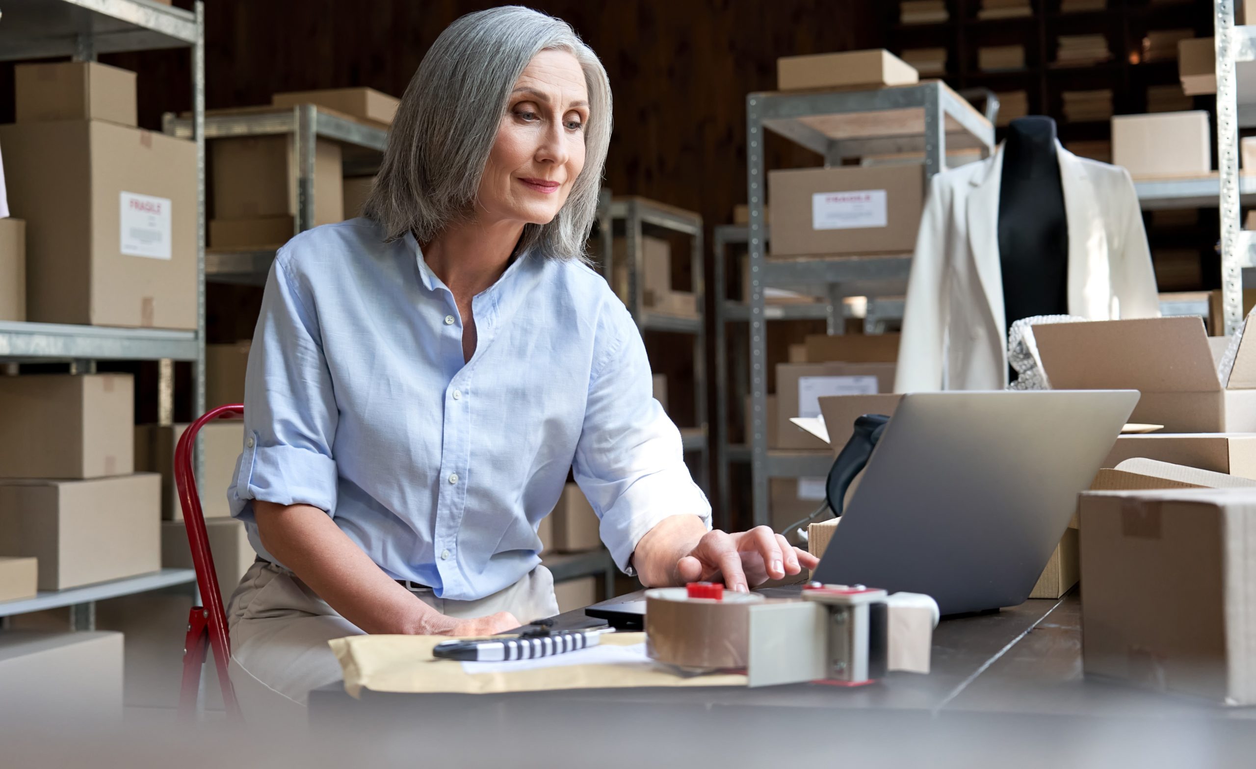 Older woman using laptop surrounded by boxes of stock, Black Friday concept