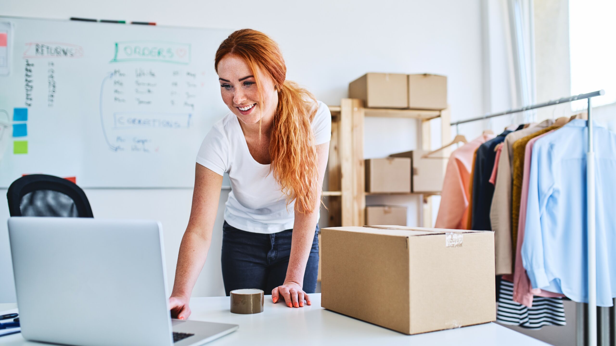 Pretty redheaded girl poised over laptop with clothes rail in background, Forward Advances concept