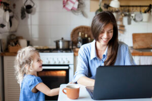 Happy young mother working at laptop in kitchen with toddler, business rates working from home concept
