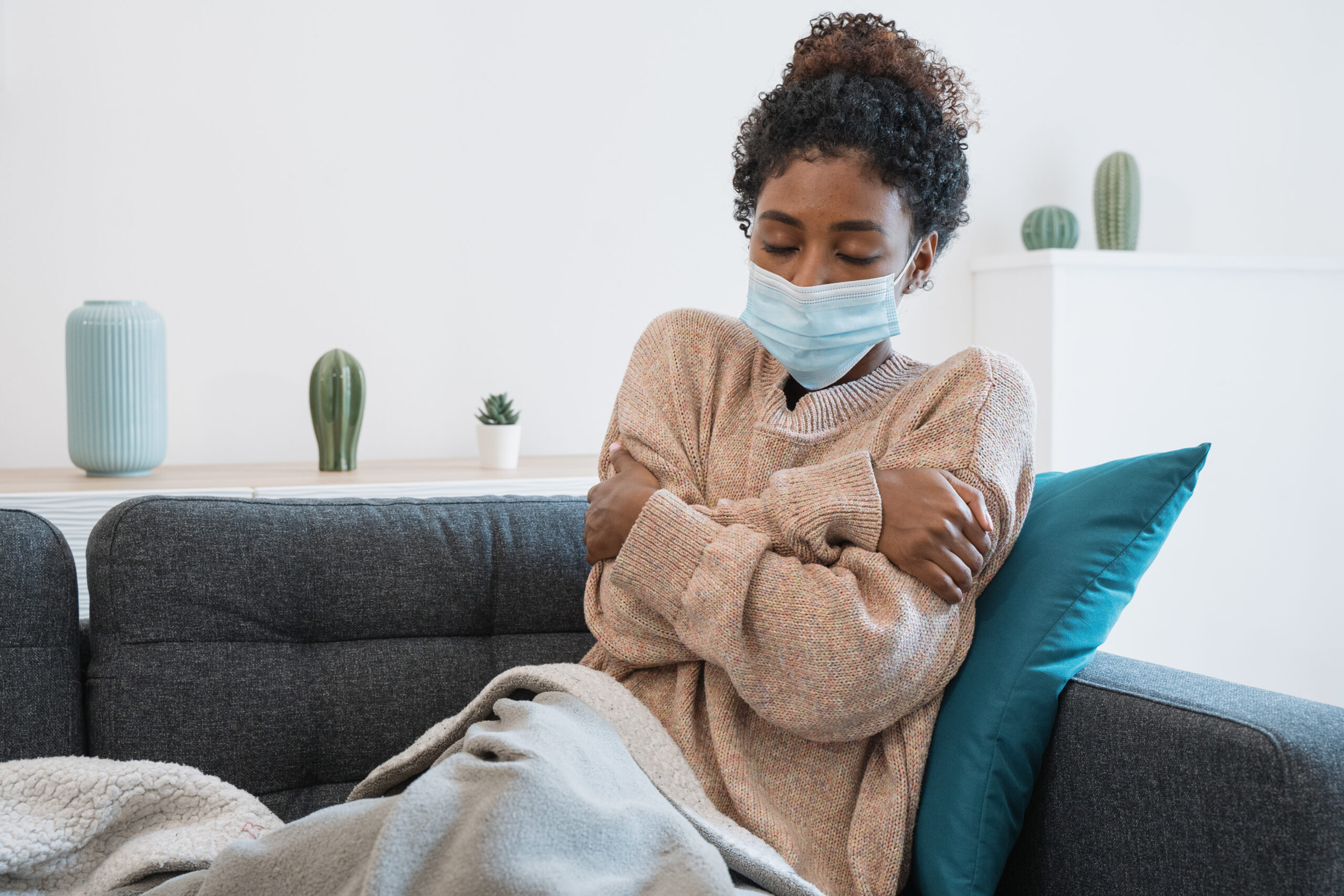 Young woman in facemask hugging herself on a sofa, staff self isolating concept