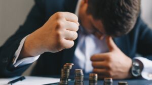 Businessman banging fist on desk behind declining piles of coins, insolvency concept
