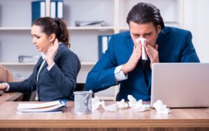 Male office worker sneezing surrounded by tissues, aghast female colleague, Covid concept