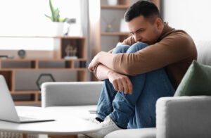 Young man hugging knees staring at open laptop in distress