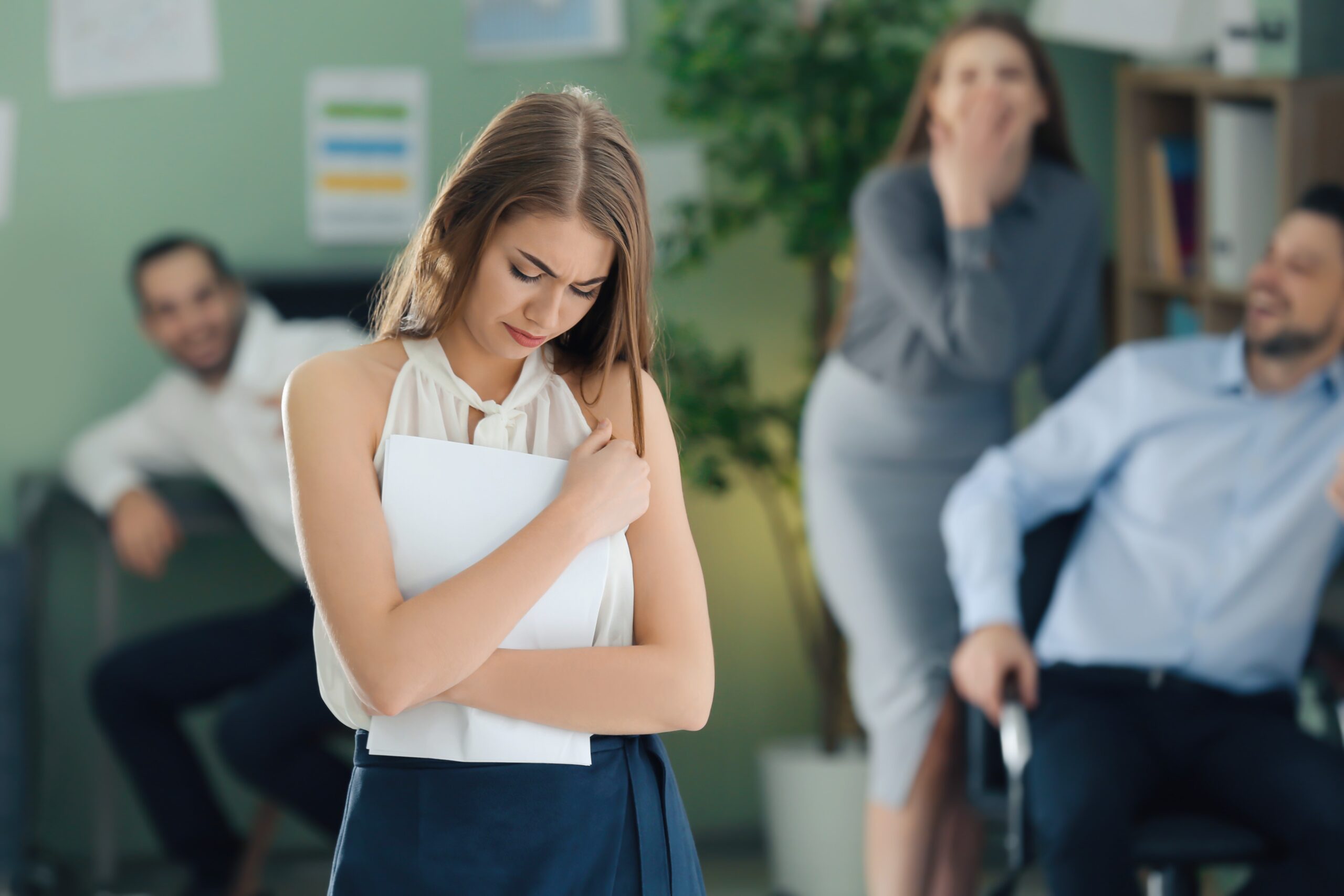 Upset young woman with jeering workmates out of focus behind her, workplace banter concept