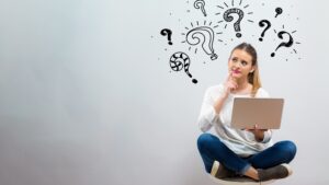 Young woman sitting cross-legged pondering with laptop surrounded by question marks, alternative business funding concept