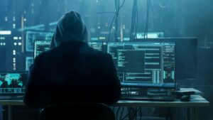 Fraud concept. Hooded hacker with his back to camera hunched over desktop PCs in dark hacker lair