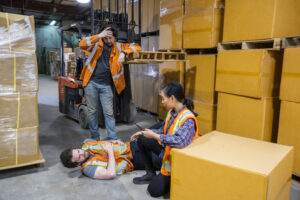 An industrial warehouse workplace safety topic. A worker injured falling or being struck by a forklift. Falls and collisions are major contributors to forklift safety. Coworkers come to his aid.