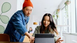 Affiliate marketing concept. Young male Asian office worker wearing red beanie hat shows co-worker something on laptop screen.