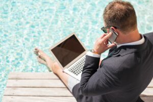 Entrepreneurs holiday concept. Suited businessman on mobile phone using laptop sitting by swimming pool with bare legs.