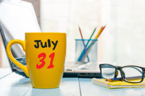 Self-employed tax concept. Yellow mug with July 31 printed on it sitting on desk beside spectacles and pen pot