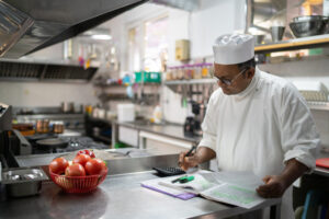 An Indian chef is writing up his daily accounts, working on the kitchen finances while standing at a counter in a commercial kitchen